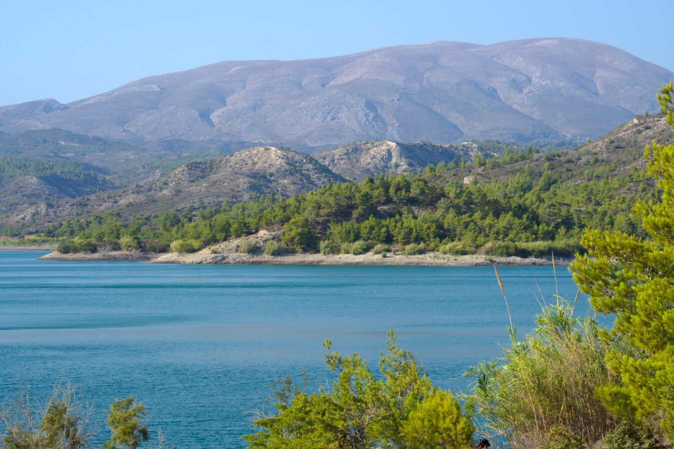Scenic morning view of the quiet turquoise freshwater reservoir at Apolakkia Dam in South Rhodes surrounded by pine trees and rolling hills.