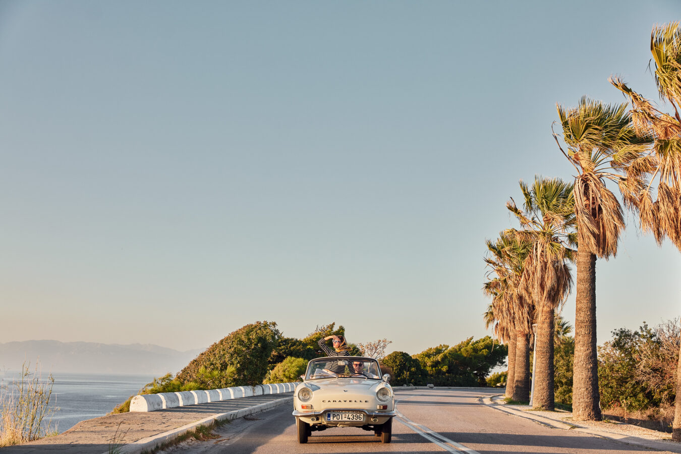 A couple in a open top car driving along on a Rhodes west coast road trip in Greece.