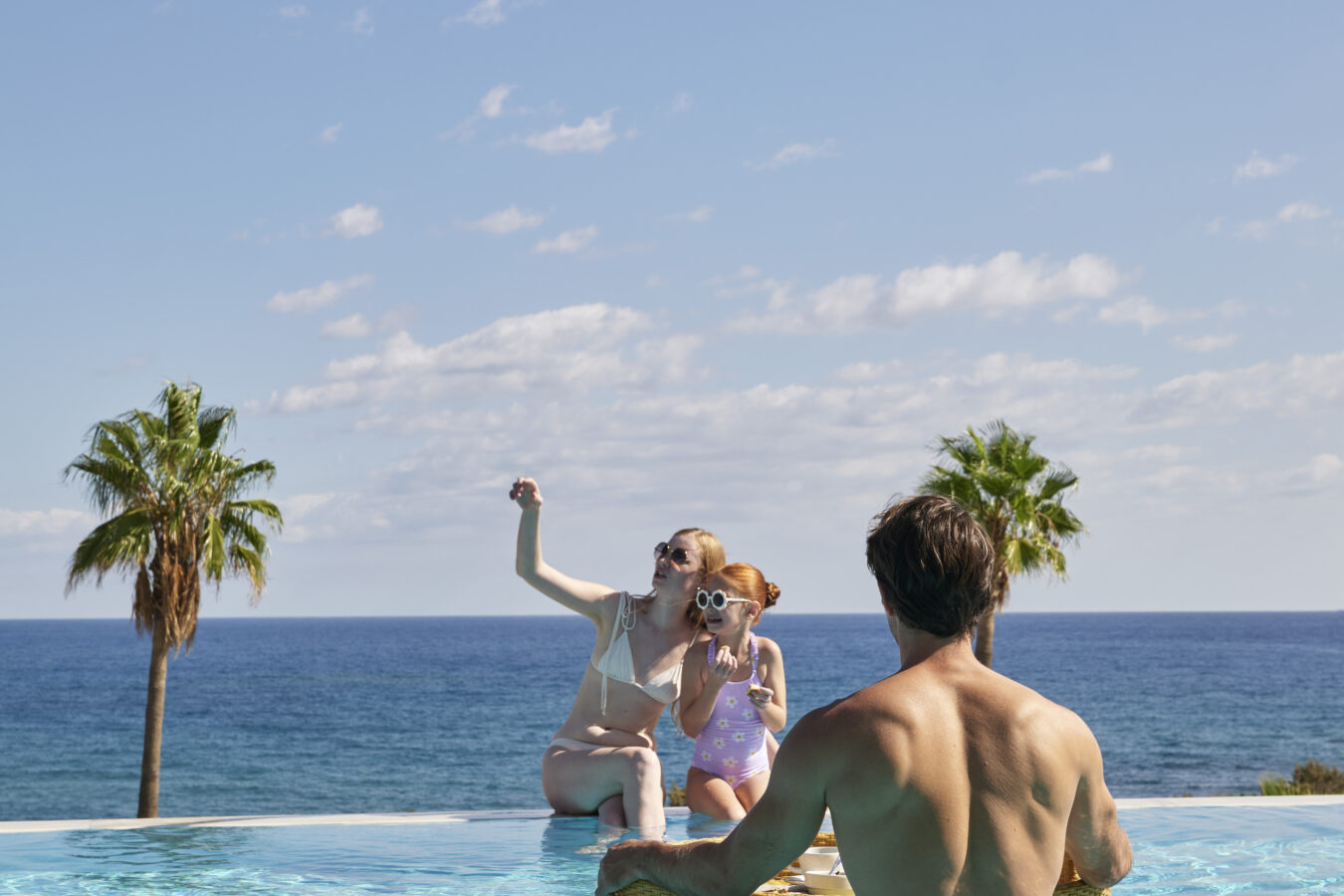 Family enjoying a floating breakfast in a private infinity pool overlooking the Aegean Sea and palm trees at a luxury resort in South Rhodes.